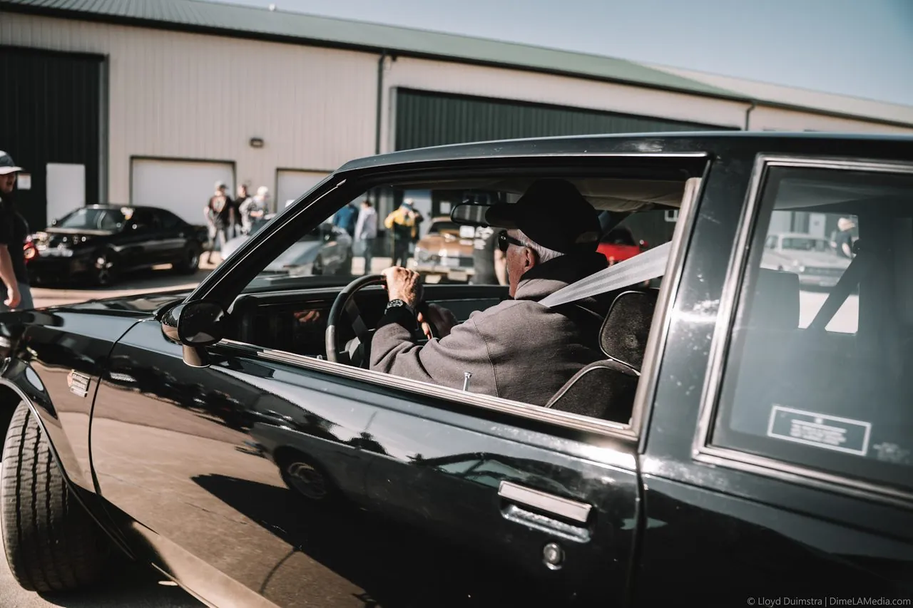 Older owner driving a black classic coupe into a South Dakota car show