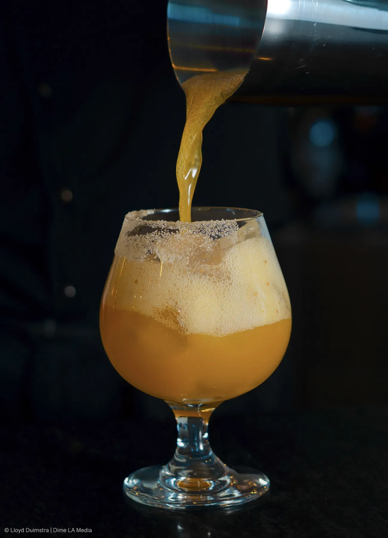 Amber cocktail with a sugared rim being poured from a shaker into a snifter glass