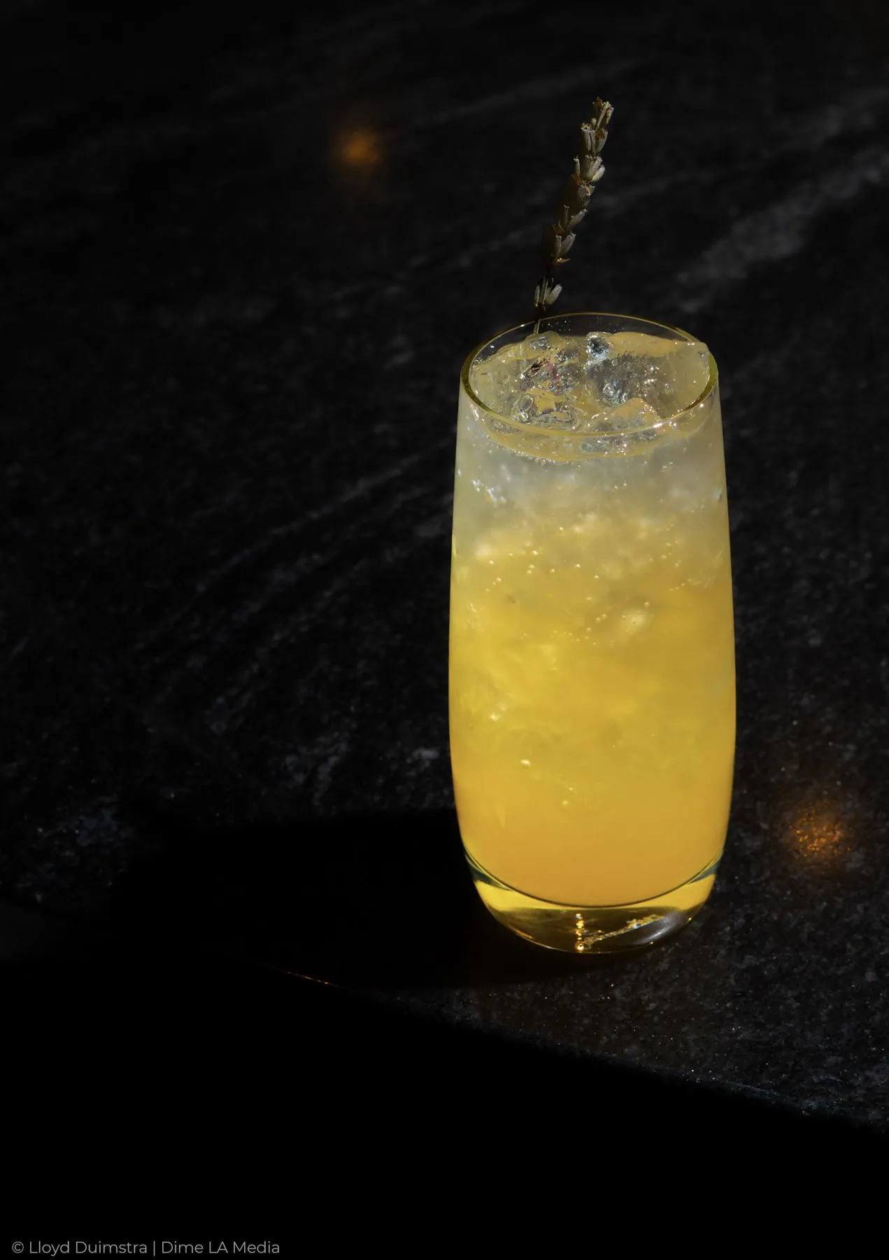 Bright yellow cocktail with a lavender sprig on dark stone