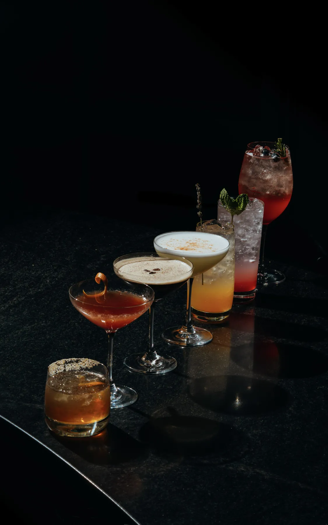 A row of cocktails receding into shadow on a black bar