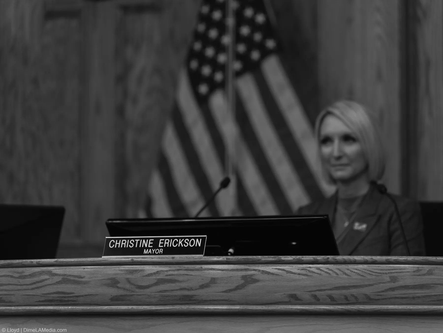 Black and white frame of the Christine Erickson mayor nameplate at the dais, the new mayor soft in the background