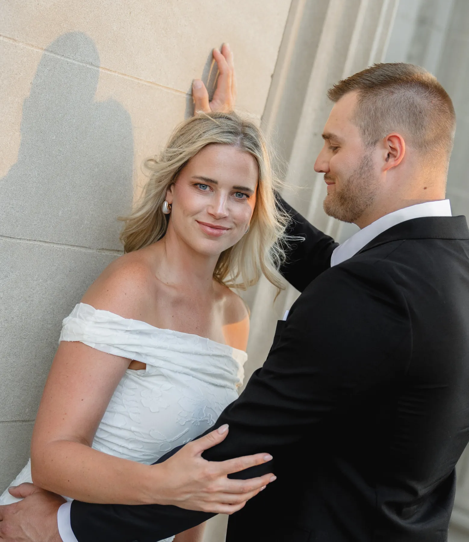 Woman in a white dress leaning against a limestone wall as her fiance leans in, downtown Sioux Falls SD