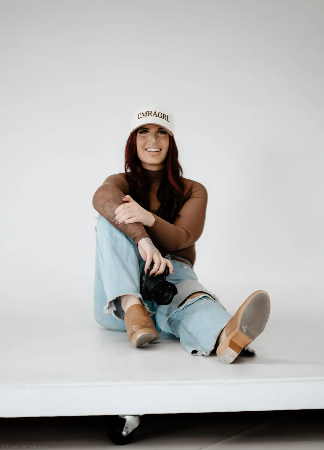 Jade sits on a studio riser with her camera resting at her knee