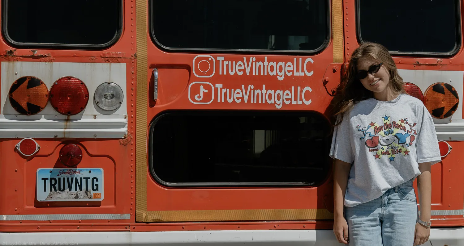 Maddie Theisen in sunglasses leans on the side of the True Vintage bus beside the TRUVNTG plate