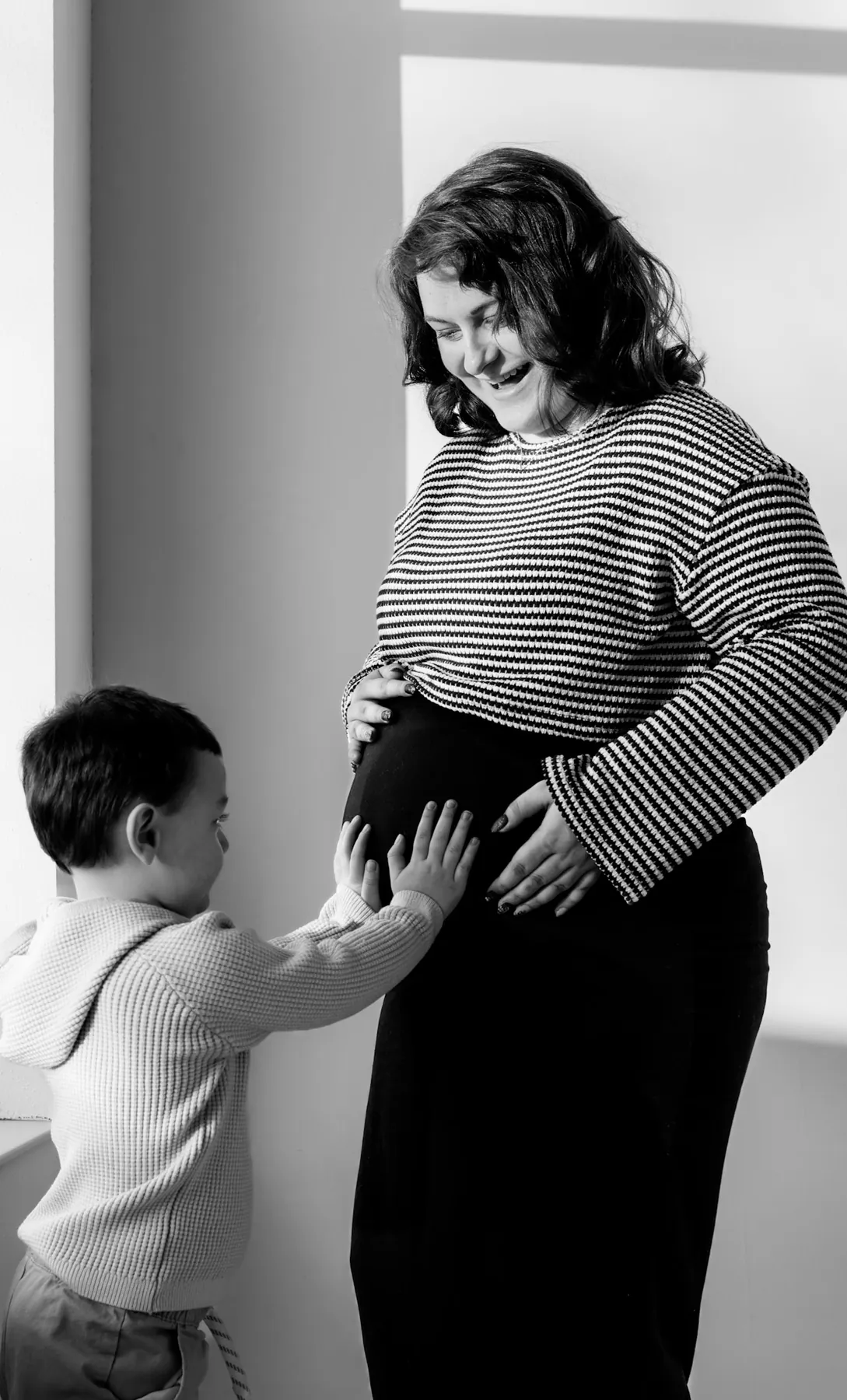 Black and white maternity portrait with a toddler reaching for mom