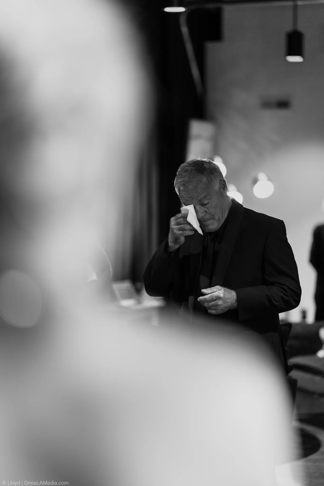 Older man in a black suit wiping his eyes, black and white, blurred foreground