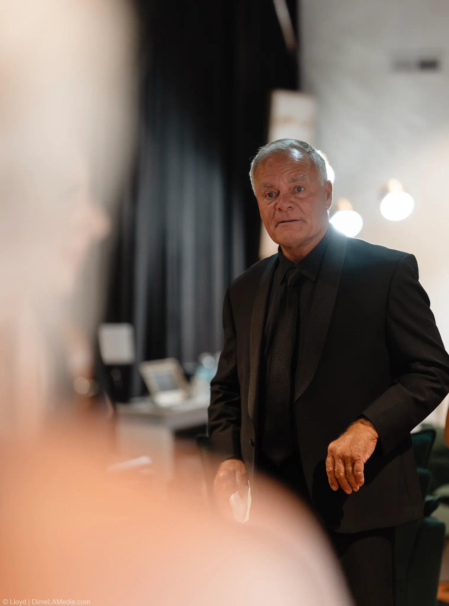 Older man in a black suit standing in a bright room, blurred foreground