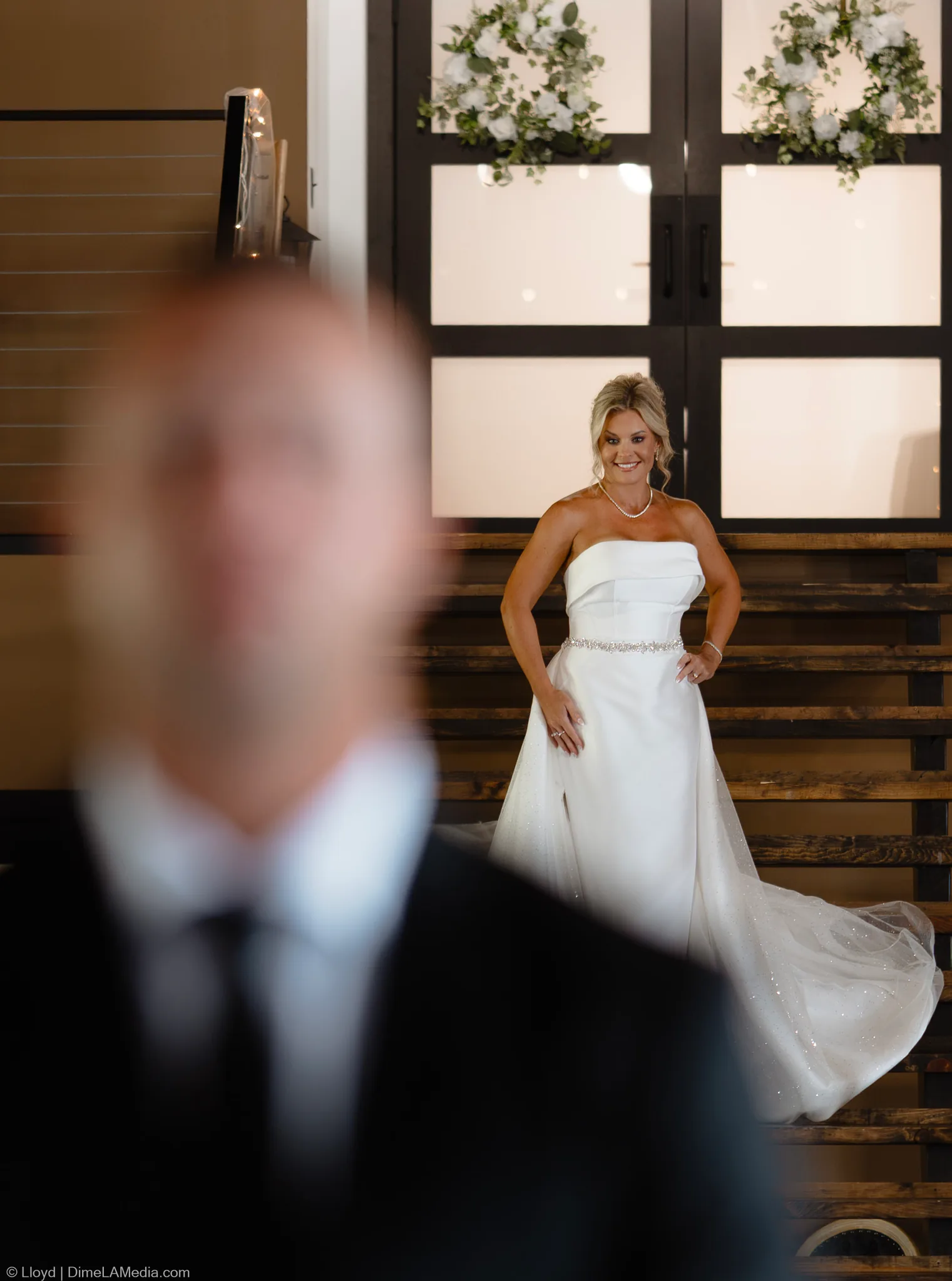 Bride in a strapless gown on wooden stairs, the groom blurred in the foreground