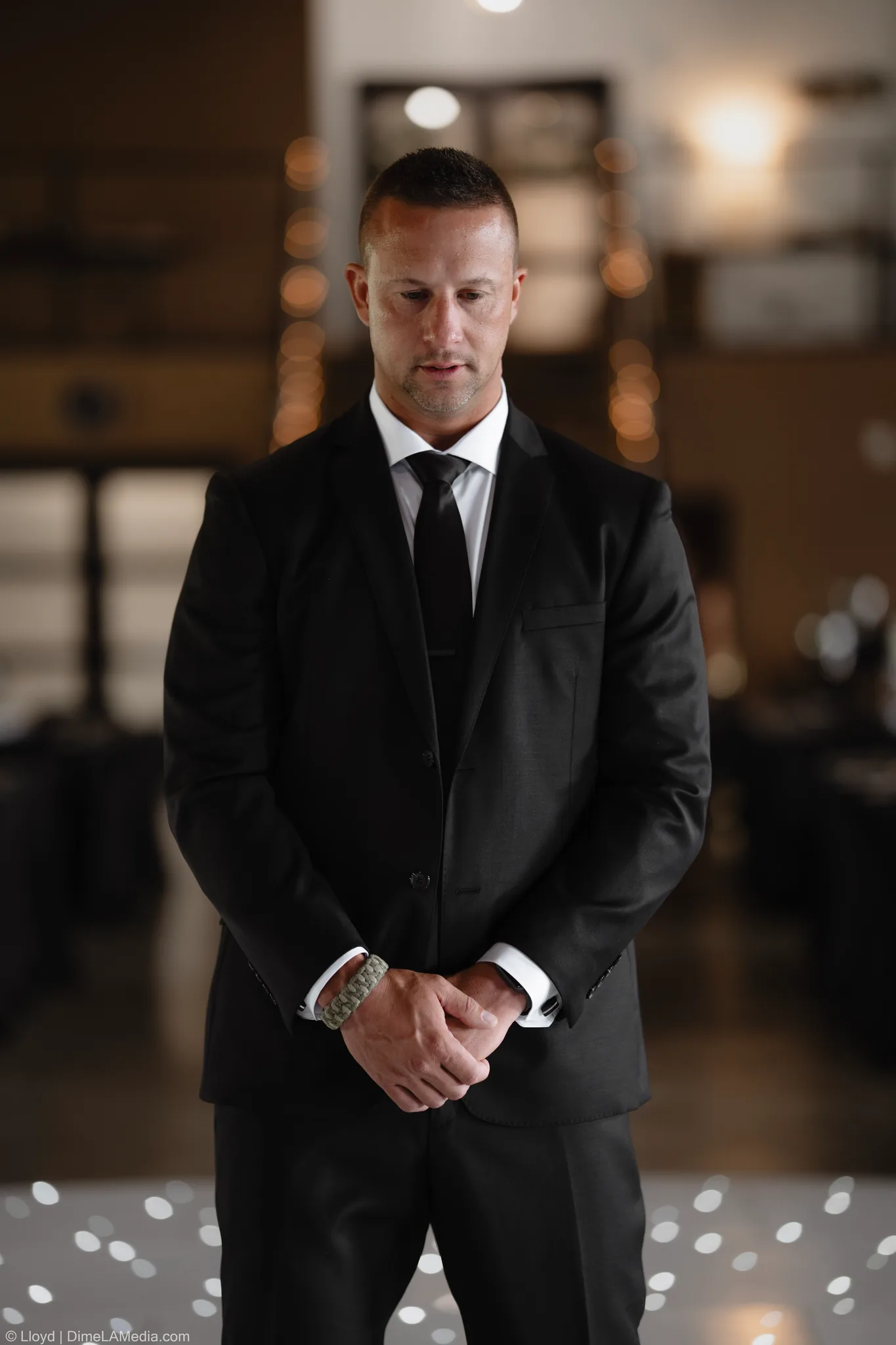 Groom in a dark suit standing with his hands clasped in a dim, warm room