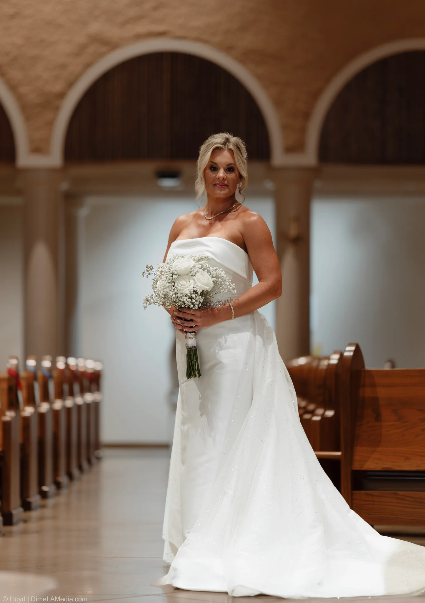 Bride in a strapless gown standing in a church aisle with her bouquet