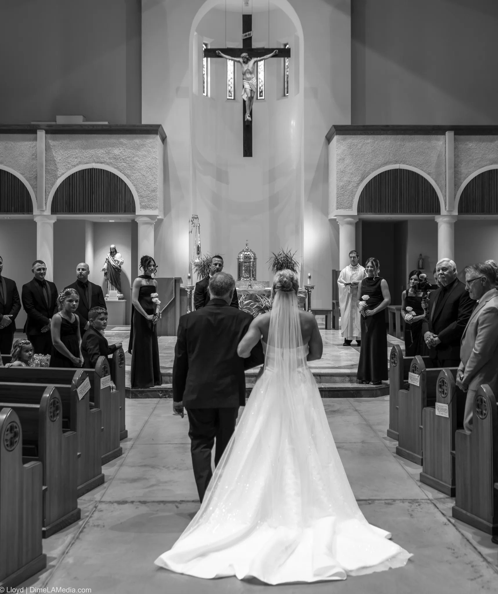 Black and white view down the aisle as the bride walks in her veil and train