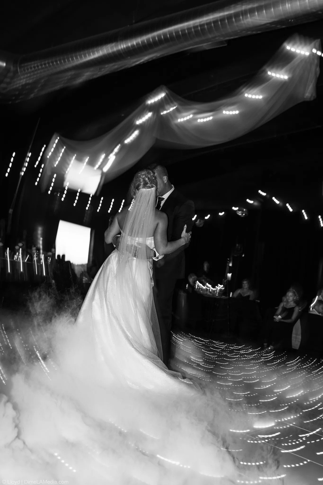 Black and white of the couple dancing through drifting fog under string lights