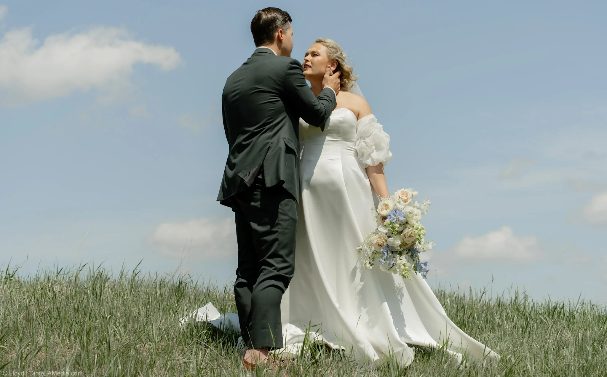 Groom holding his bride's face on a grass hilltop under towering summer clouds