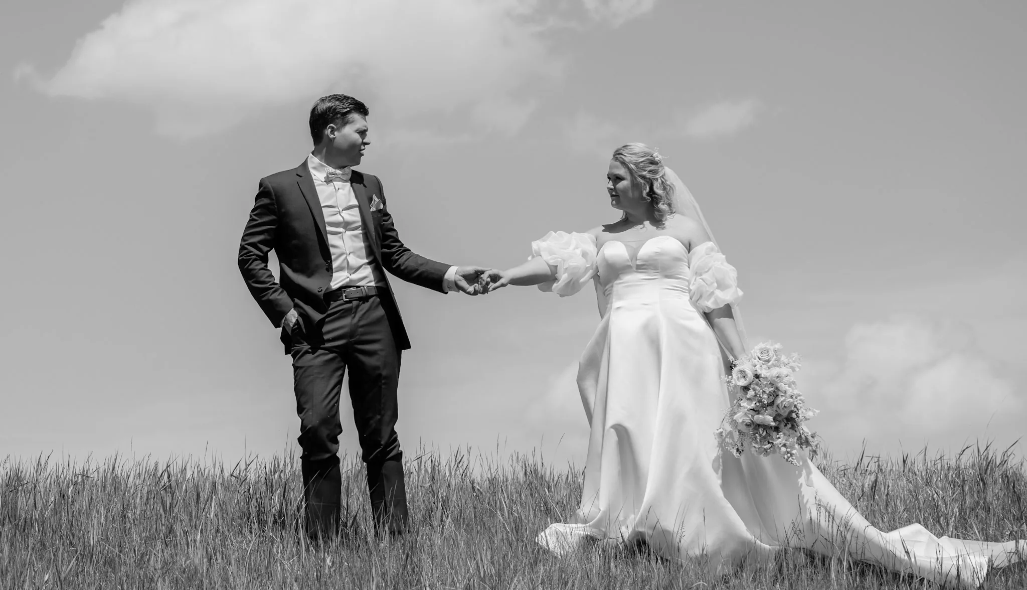 Black and white portrait of a bride and groom holding hands apart in tall prairie grass