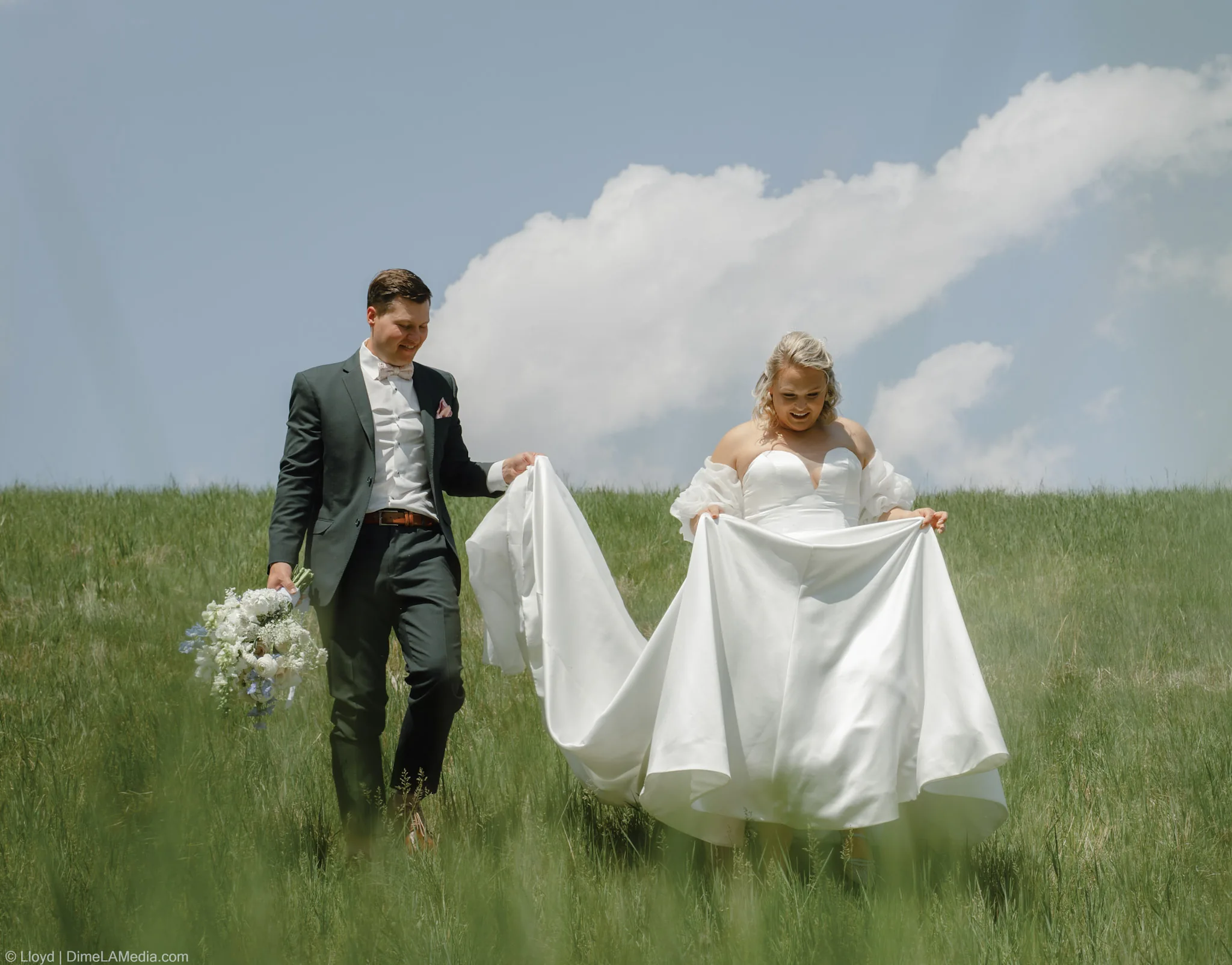 Groom carrying his bride's train and bouquet as the two walk through tall grass barefoot