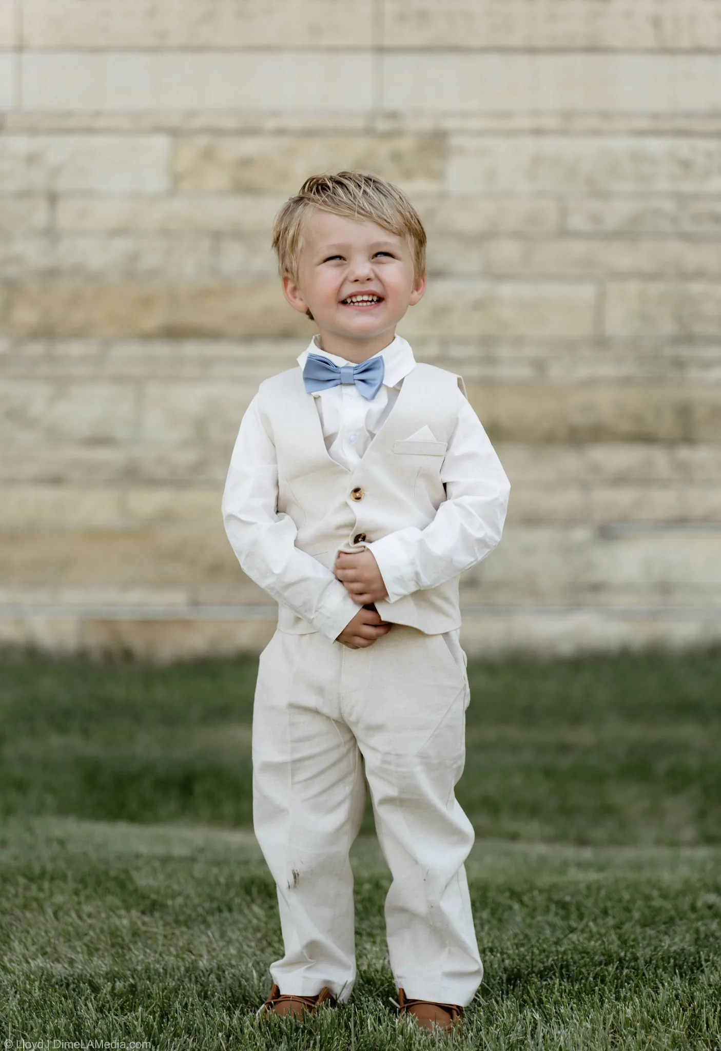 Ring bearer in a cream linen suit and blue bow tie grinning at the camera before the ceremony
