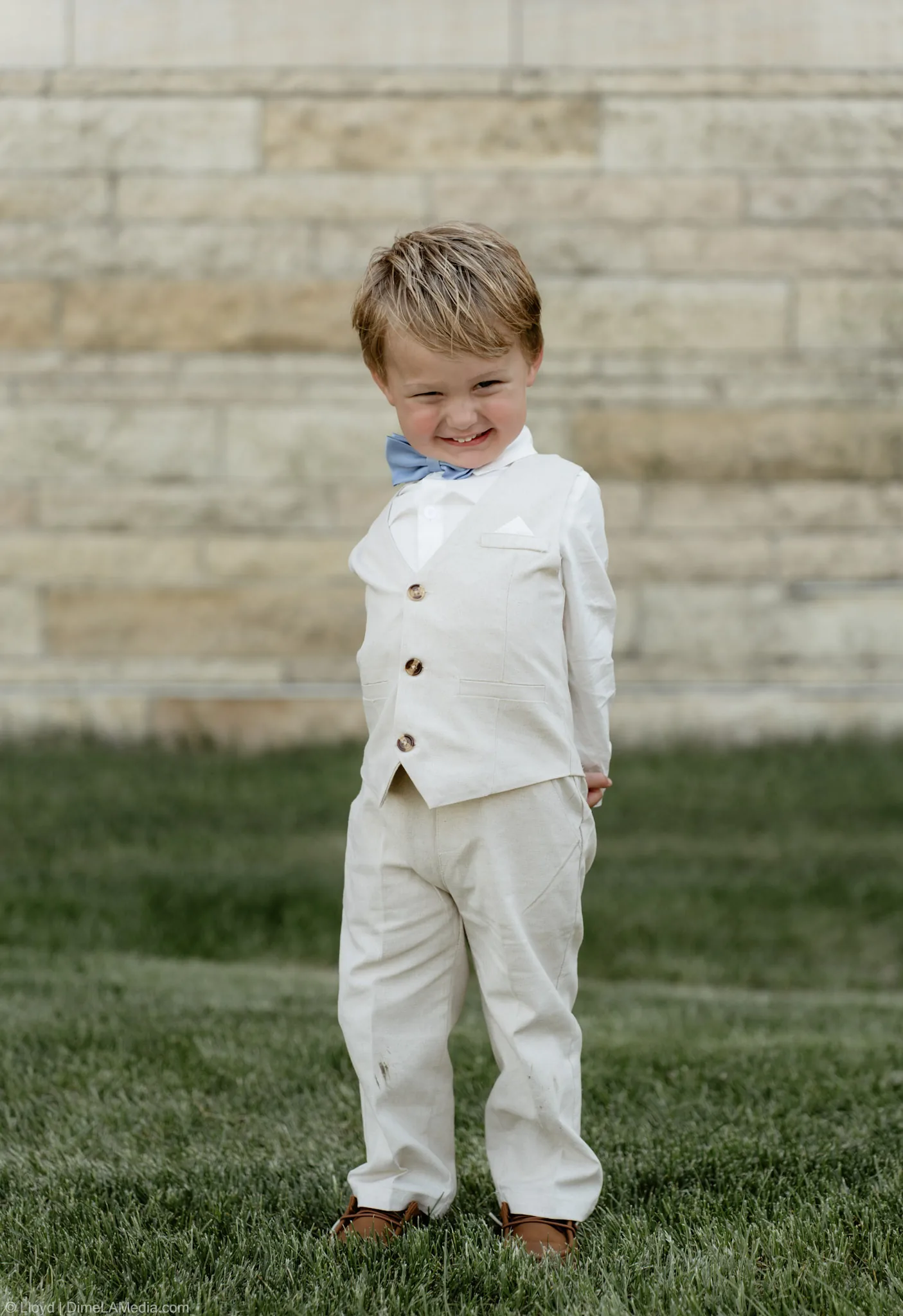 Ring bearer squinting into the sun in his cream linen suit and blue bow tie