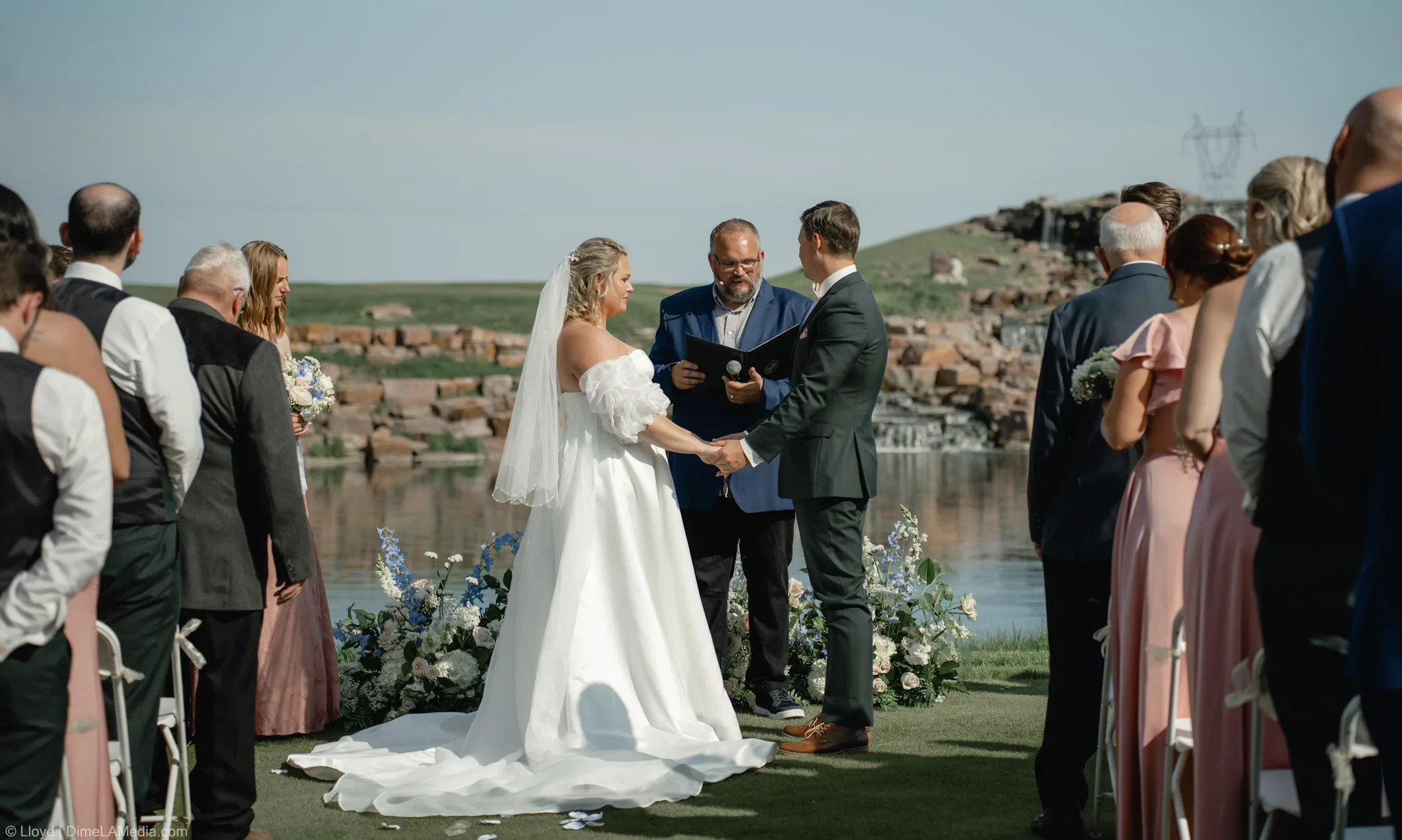 Wide view of an outdoor wedding ceremony beside a quarry waterfall with guests seated on the lawn
