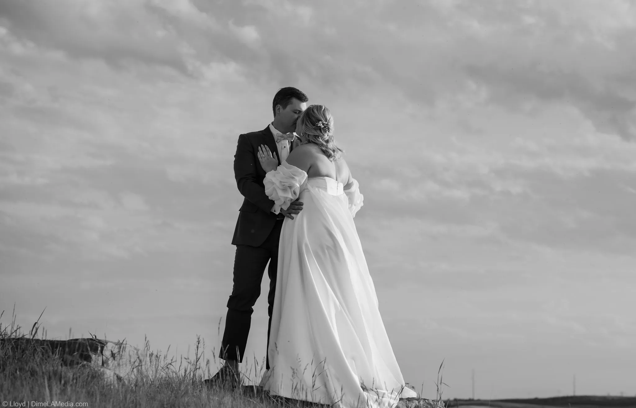 Black and white portrait of a bride and groom embracing on a hilltop under a dramatic sky